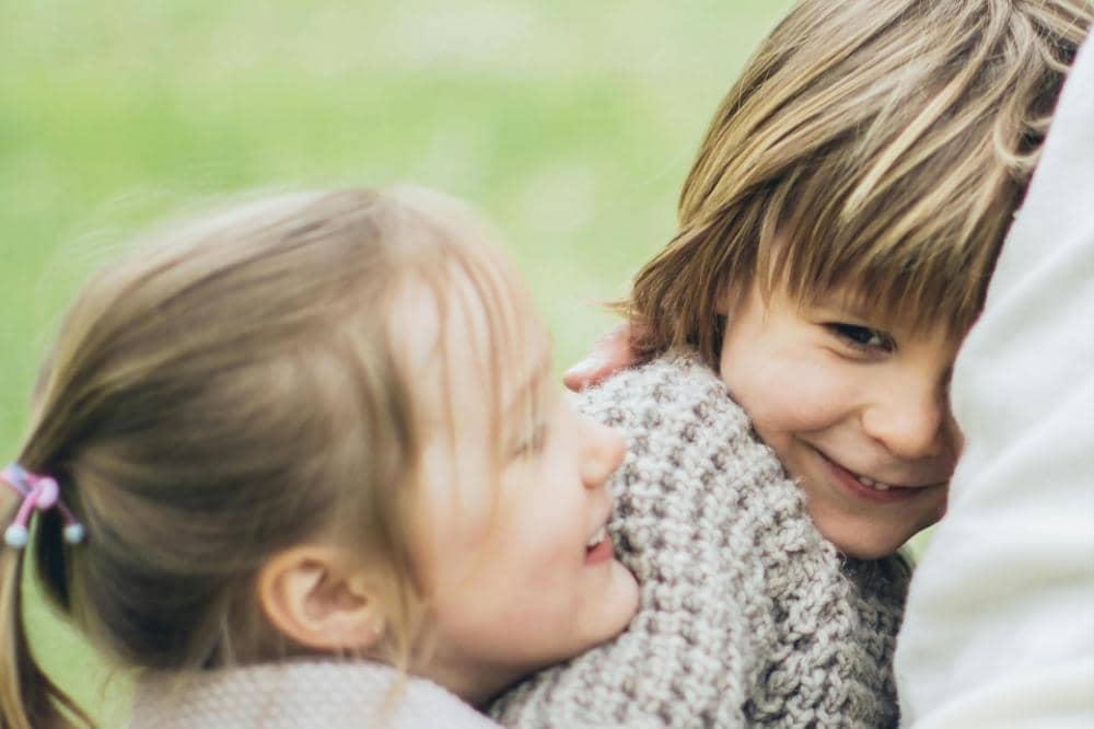 Dua anak kecil tersenyum bahagia sambil berpelukan di luar ruangan dengan latar belakang hijau yang lembut.