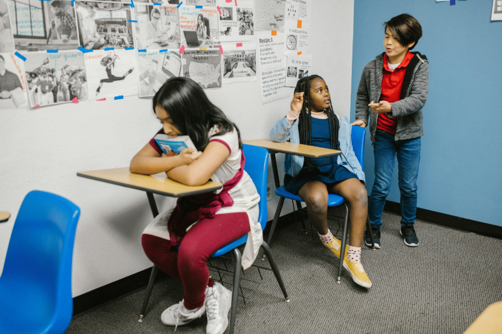 Tiga anak di ruang kelas, satu anak duduk membaca buku sementara dua lainnya berbincang