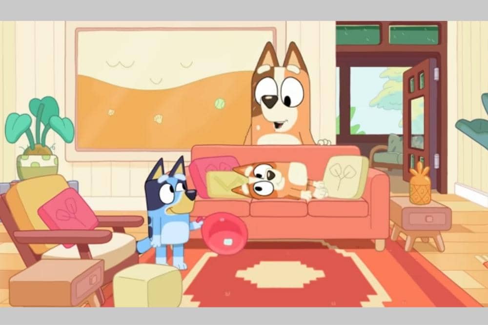 Bluey, Bingo, dan Chilli bermain bola merah di ruang tamu dengan sofa oranye dan dekorasi hangat.