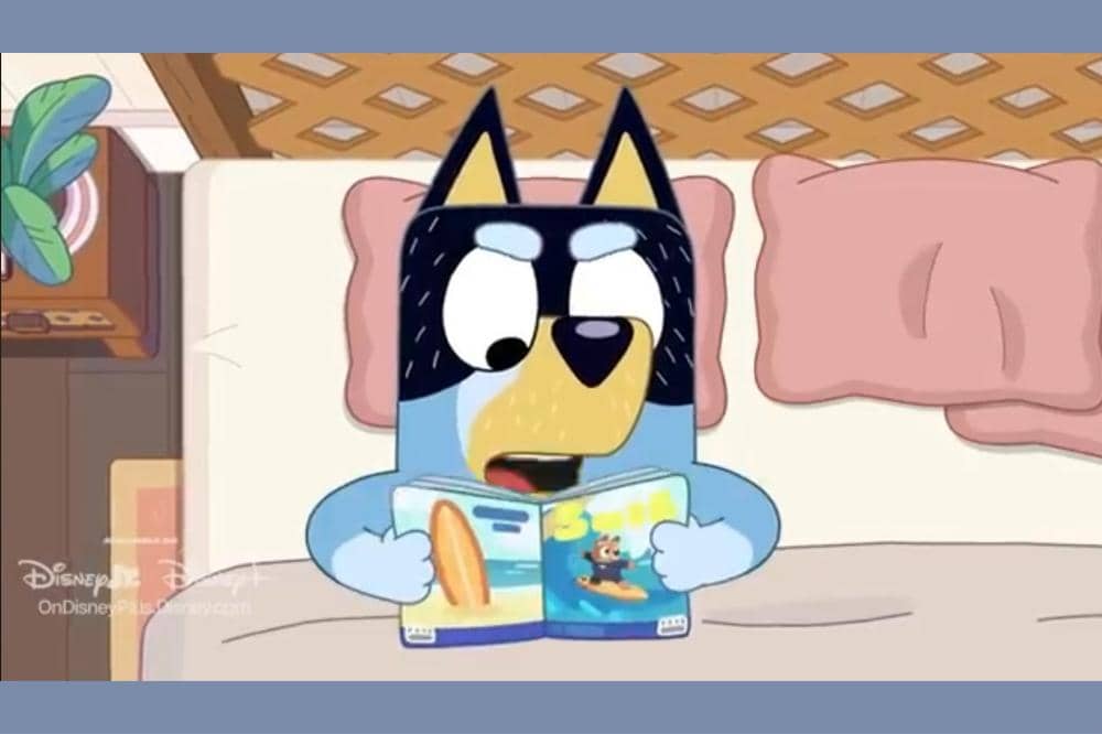 Karakter Bluey sedang duduk di tempat tidur sambil membaca buku bergambar di kamar dengan bantal merah muda.