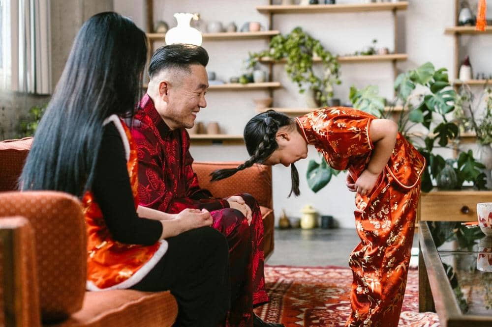 Seorang anak perempuan mengenakan pakaian tradisional merah membungkuk memberi hormat kepada orangtua yang duduk di ruang tamu hangat dan penuh tanaman.