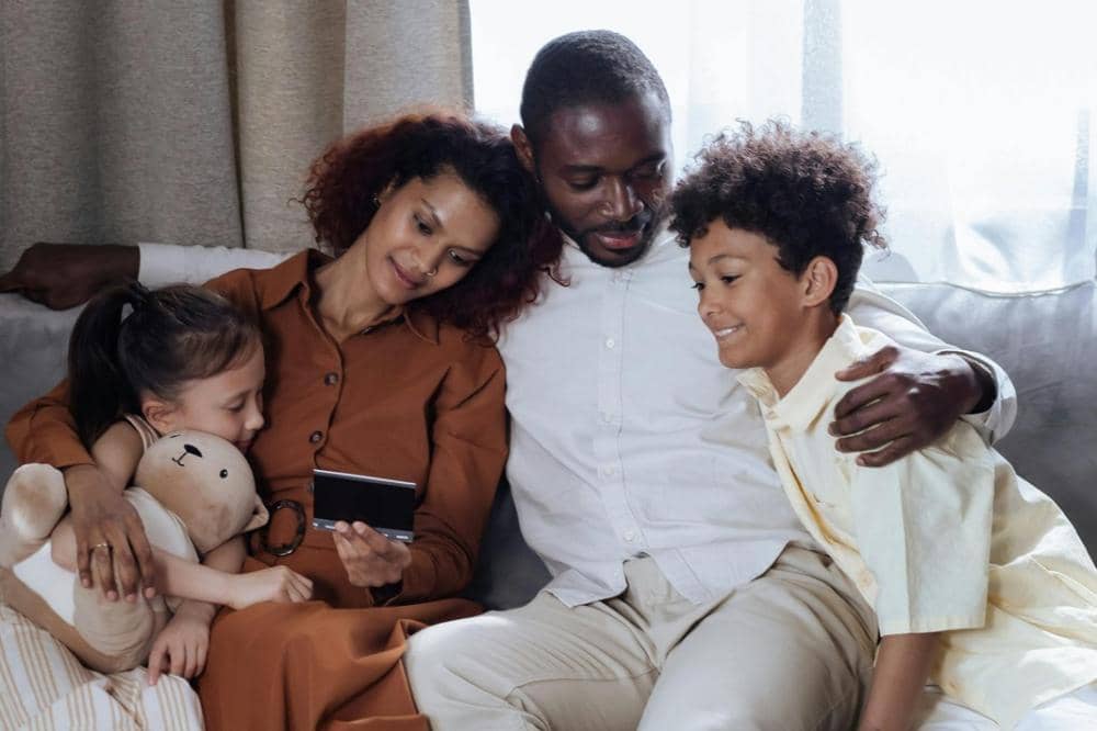 Keluarga duduk bersama di sofa sambil menonton sesuatu di ponsel, anak kecil memeluk boneka dan tersenyum bahagia.