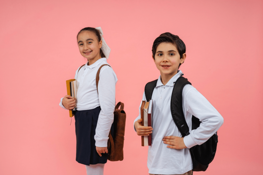 Dua anak sekolah mengenakan seragam putih dan membawa tas serta buku di tangan