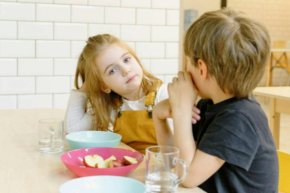 Dua anak duduk di meja makan dengan mangkuk berisi potongan apel dan gelas air di depan mereka di ruangan terang dengan dinding ubin putih.