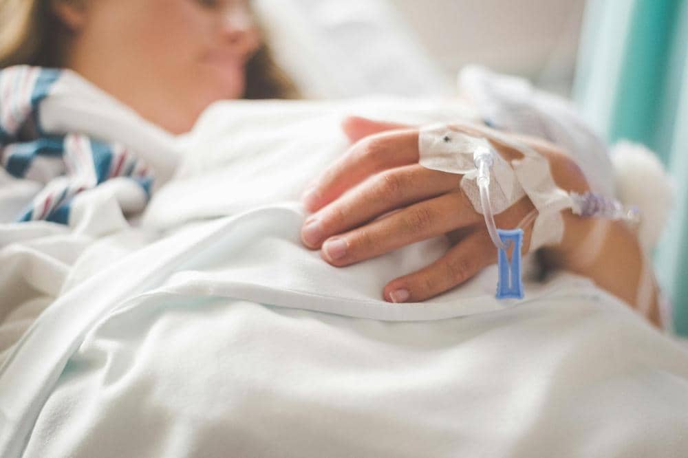 Seorang pasien berbaring di tempat tidur rumah sakit dengan infus terpasang di tangan, mengenakan selimut putih dan tampak beristirahat.