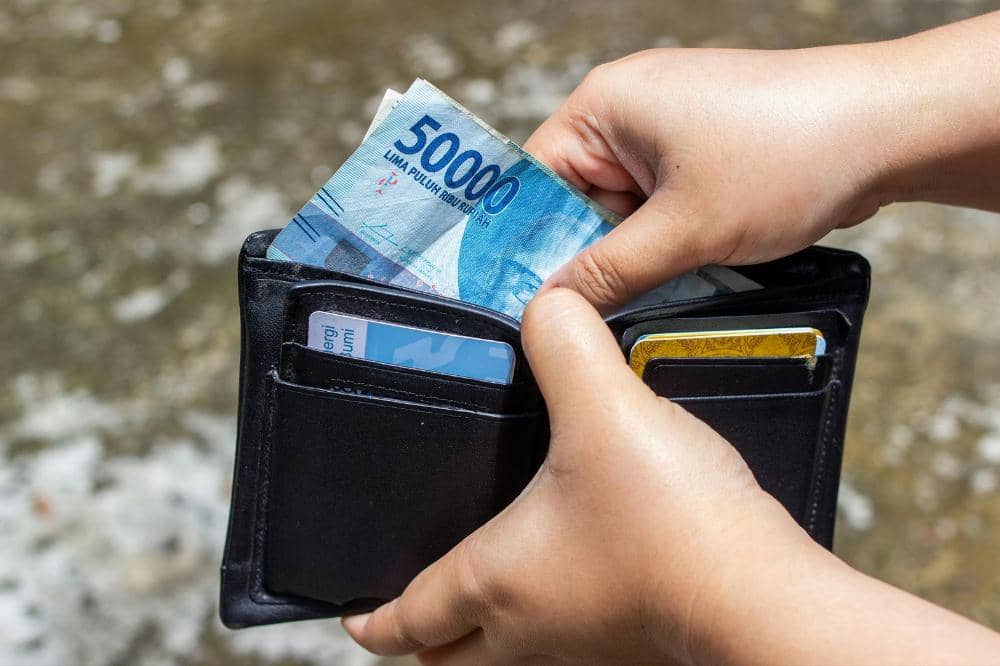 Seseorang memegang dompet hitam berisi uang kertas lima puluh ribu rupiah dan beberapa kartu di dalamnya.