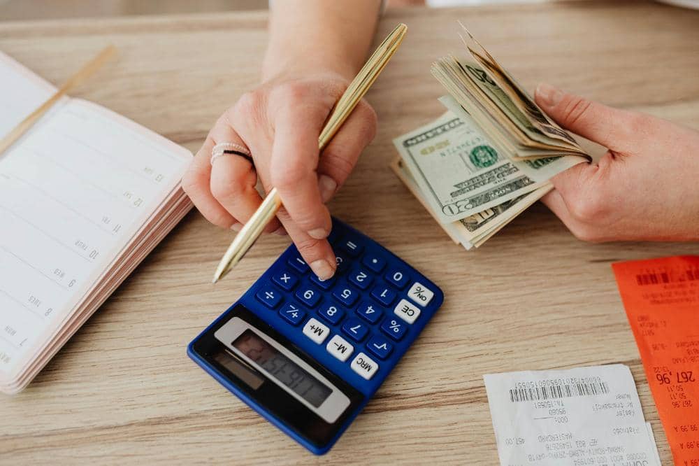 Seseorang menghitung uang tunai dolar AS sambil menggunakan kalkulator biru dan mencatat di buku catatan di atas meja kayu.