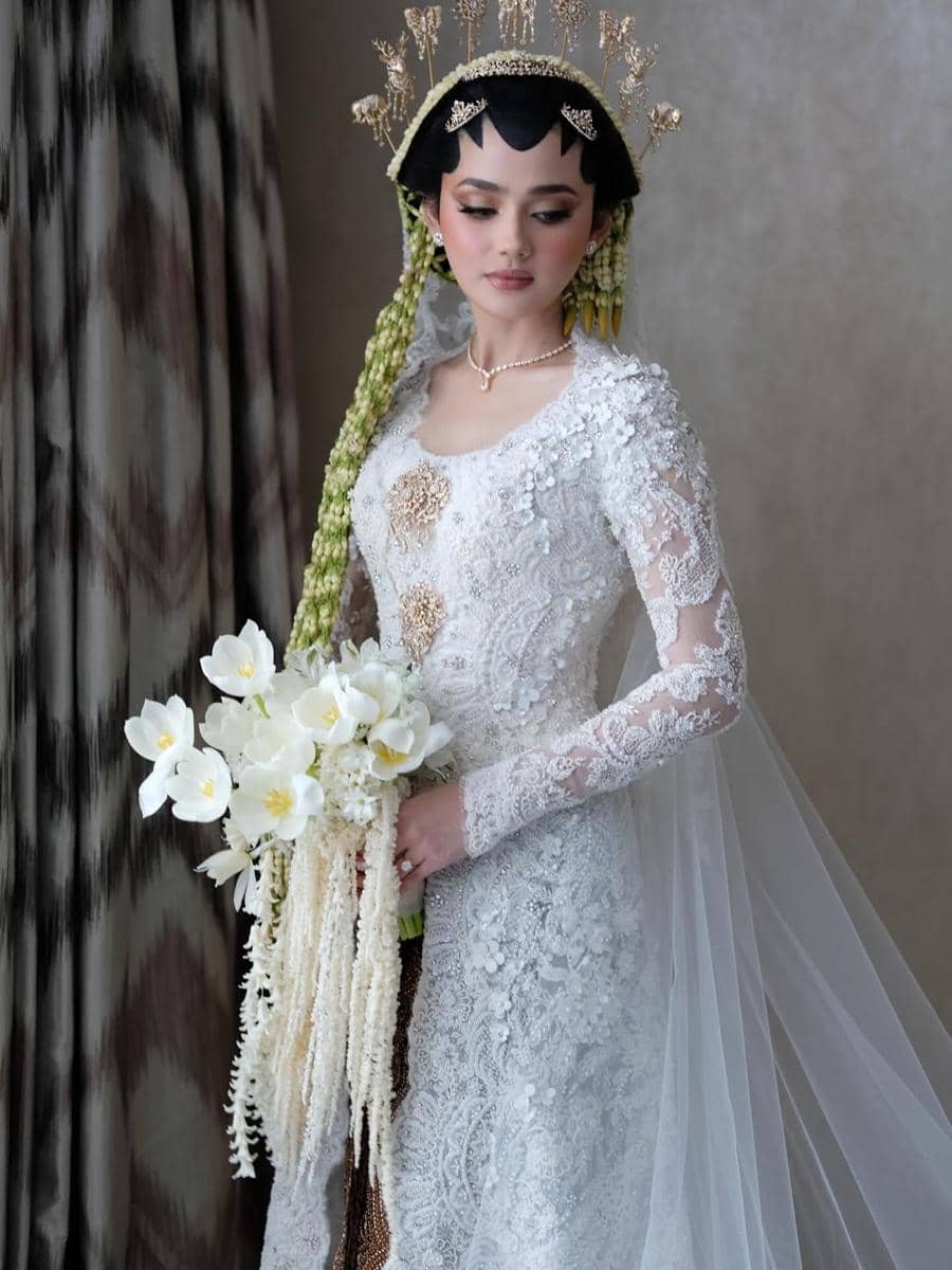 Syifa Hadju mengenakan kebaya putih berhias renda dan siger Sunda, memegang buket bunga putih dengan selendang panjang.