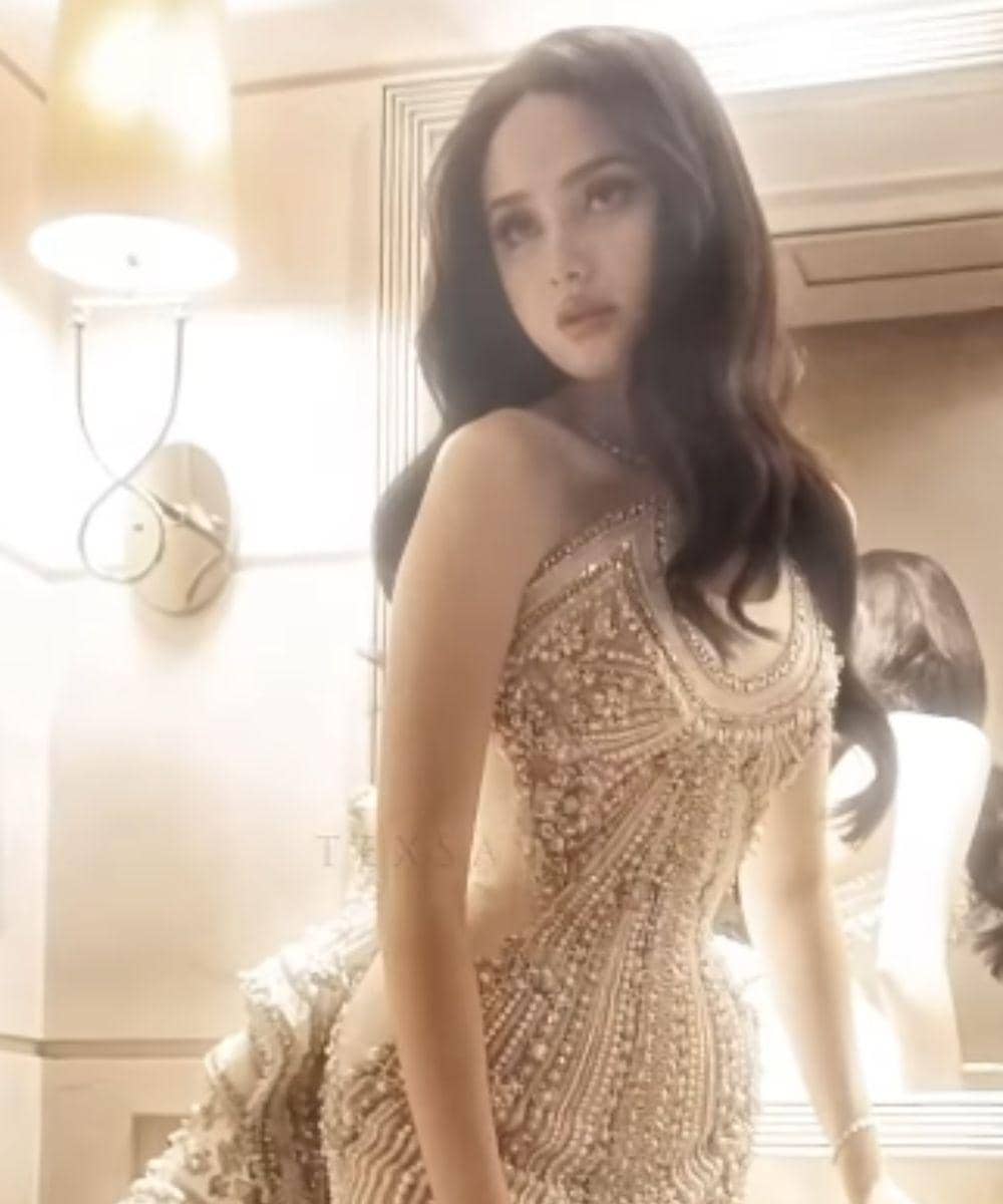 Syifa Hadju mengenakan gaun mewah berwarna emas dengan detail manik-manik rumit berdiri di depan cermin dalam pencahayaan hangat.