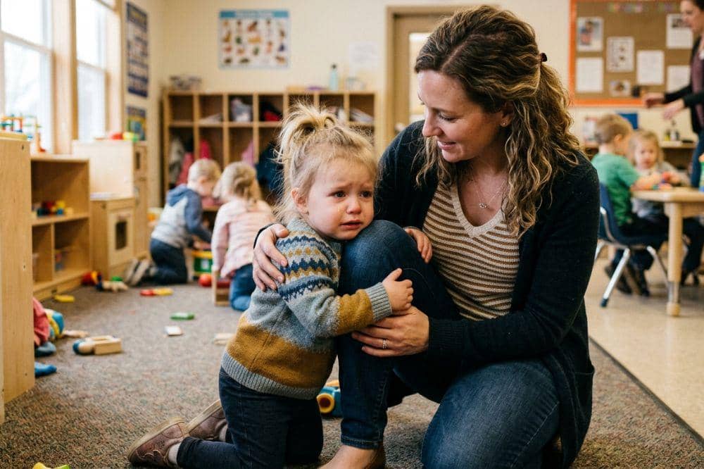 Seorang anak kecil menangis di ruang kelas taman kanak-kanak sambil dipeluk oleh seorang perempuan dewasa yang menenangkannya.