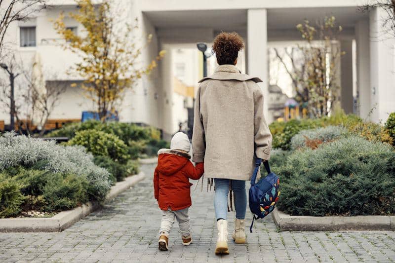 Seorang wanita berjalan sambil menggandeng tangan anak kecil yang mengenakan jaket merah di area perumahan dengan taman hijau.