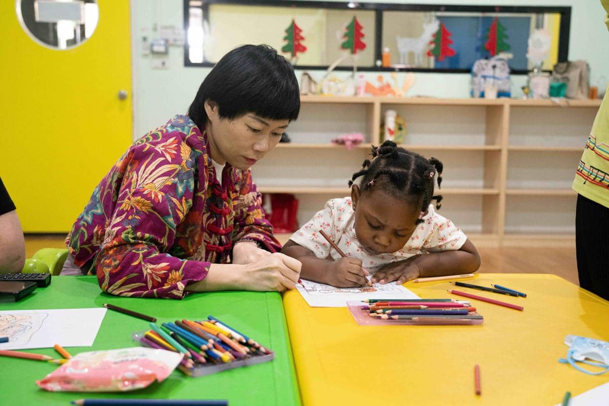 Guru membantu anak kecil mewarnai gambar di meja kelas dengan pensil warna di ruang belajar yang cerah dan penuh dekorasi.