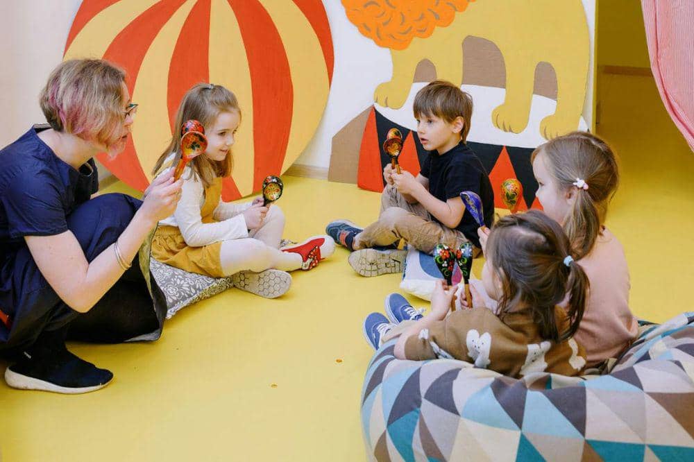 Seorang guru duduk melingkar bersama anak-anak di ruang bermain berwarna cerah sambil memegang alat musik marakas kayu.