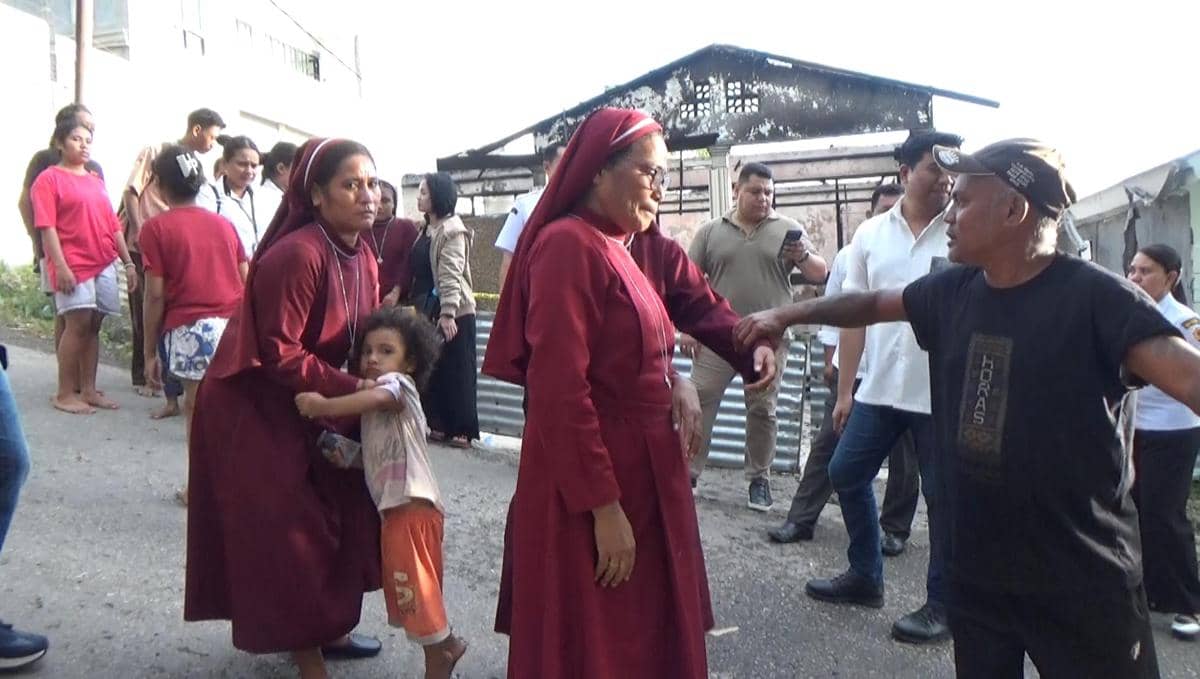 Para suster dan anak-anak panti asuhan Katolik berdiri di luar bangunan yang hangus terbakar, tampak berinteraksi dengan warga sekitar.