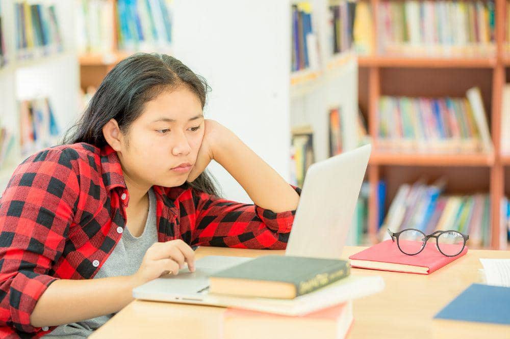 Seorang mahasiswa mengenakan kemeja kotak-kotak merah hitam tampak lelah saat belajar di depan laptop di perpustakaan dengan tumpukan buku.
