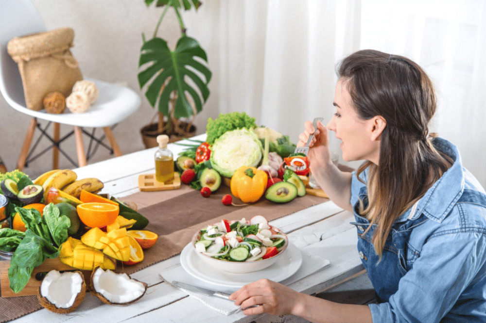 Seorang wanita duduk di meja makan menikmati salad segar dengan berbagai buah dan sayuran berwarna-warni di sekitarnya.