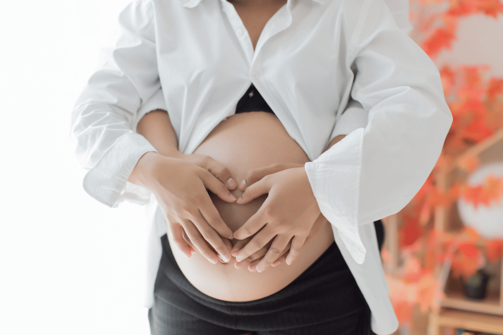 Pasangan membuat bentuk hati dengan tangan di atas perut wanita hamil yang mengenakan kemeja putih terbuka dan celana hitam.
