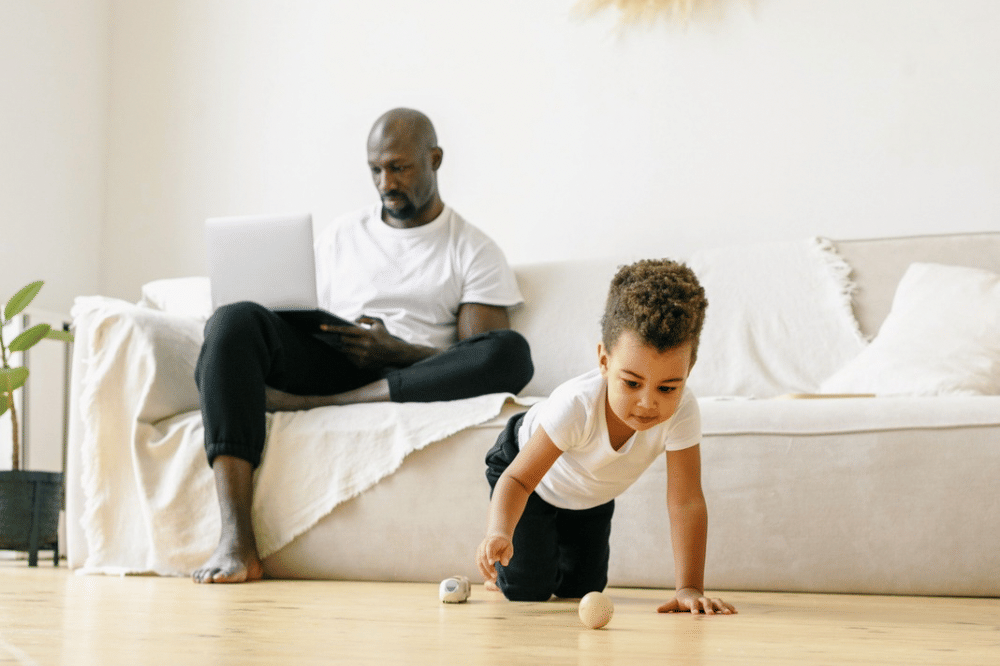 Papa bekerja di atas sofa dan sang anak bermain di lantai