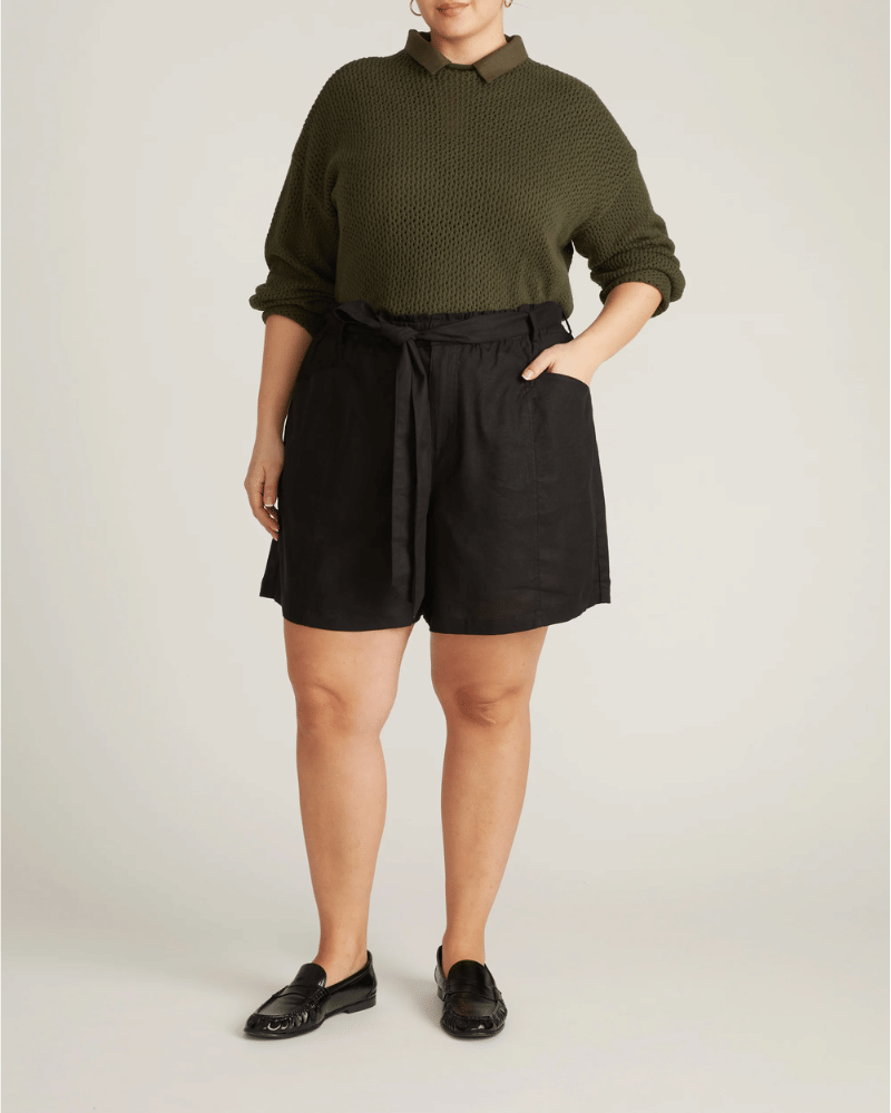 Seorang wanita mengenakan sweater rajut hijau zaitun dengan kerah dan celana pendek hitam berikat, dipadukan dengan sepatu loafer hitam.