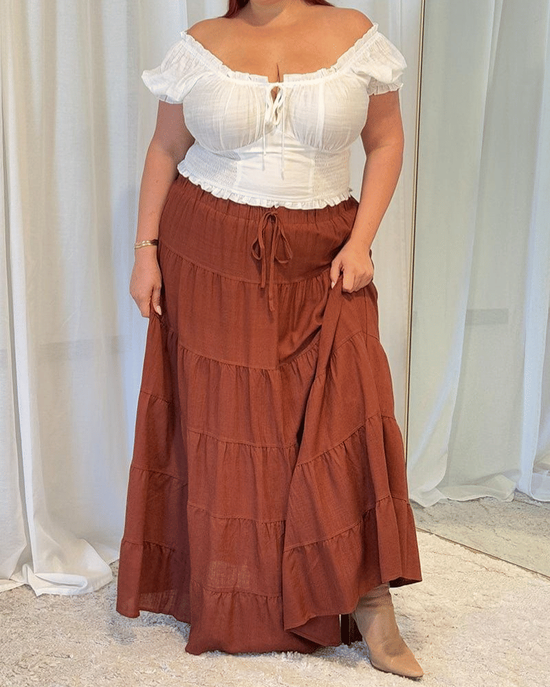 Seorang wanita mengenakan blus putih model off shoulder dengan rok panjang berwarna cokelat bata dan sepatu bot krem di ruangan berpadu tirai putih.