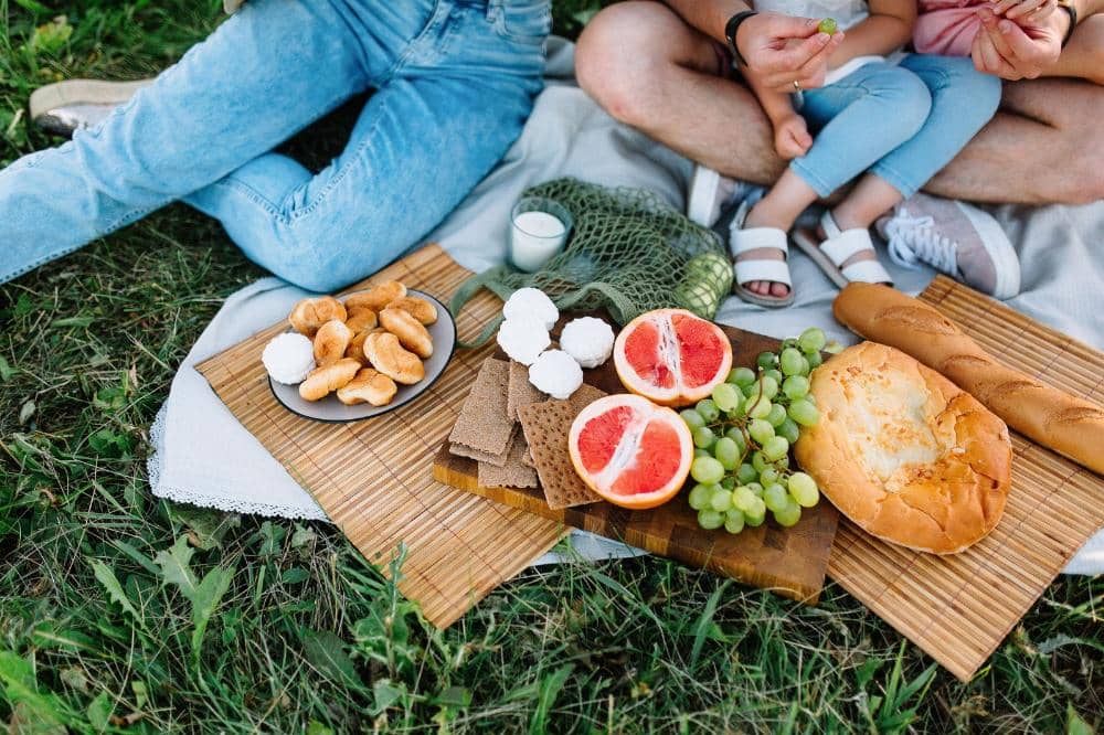 Piknik di taman dengan makanan ringan seperti roti, anggur hijau, jeruk bali, dan kue yang tersaji di atas karpet piknik