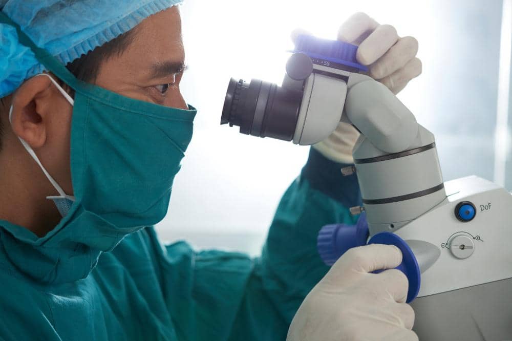 Tenaga medis mengenakan pakaian bedah dan masker sedang mengamati melalui mikroskop