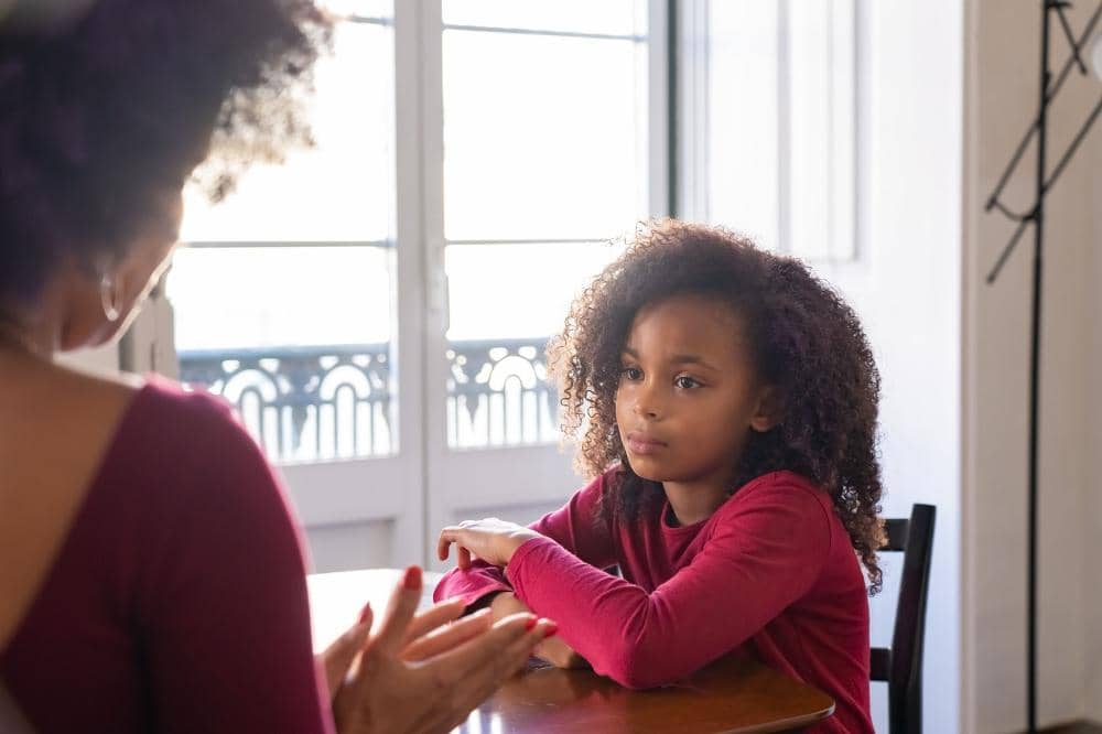 Seorang anak perempuan duduk di meja sambil mendengarkan orang Mama yang berbicara